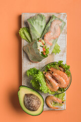 Spring rice paper roll with shrimp, avocado, paprika, lettuce and cottage cheese on a wooden white Board on an orange background. Vegan food.