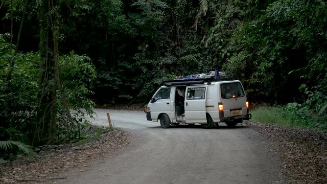 Old camper van on a dirt jungle road. Surf board on top, huge dense jungle, tropical climate