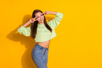 Photo of attractive pretty funny lady good mood arms showing v-sign symbols near eyes flirty wear cropped sweatshirt green sweater jeans isolated vivid bright yellow color background