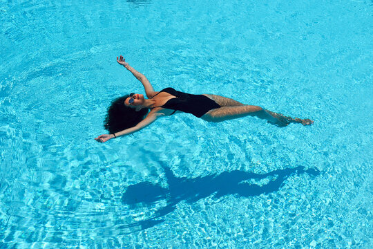 Young beautiful sexy slim tanned caucasian woman with brunette hair in black swimwear relaxing and lying in blue water of swimming pool at tropical resort. Vacation concept. Top poinit of view