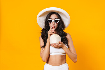 Image of pleased young woman in swimsuit and hat drinking coconut water