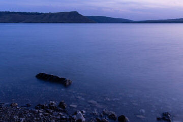 landscape evening long exposure river shore line with soft focus stone in shallow water foreground and hill land background horizon space twilight lighting after sunset
