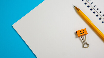 Blank Paper Notepad with Pencil and Smile Binder Clip on Blue Background. Spiral notebook on table. Business, office supplies or education concept