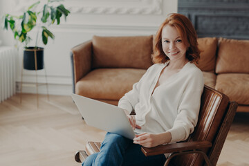 Indoor shot of happy redhead woman freelancer sits at home interior, uses modern laptop computer for distance work, enjoys online communication and free internet connection, waits for answer