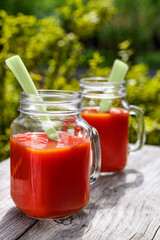 Two glasses of tomato juice with parsley and celery decorations, stand in the open air on wooden boards, the rays of the sun shine, shallow depth of field, selective focus. The concept of natural