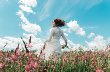 Back view portrait of young woman in motion in a long white dress walking with a wild flower bouquet in a blossoming flower field holding dress with hand