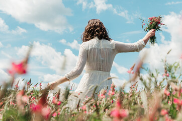 Back view of woman in long white dress walking with a wildflower bouquet in pink flower field