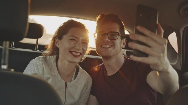 Young male blogger making selfie with charming asian girl on phone smartphone in back seat of car at sunset. Friends smiling on camera. Handsome man in glasses travelling by car. Concept of roadtrip