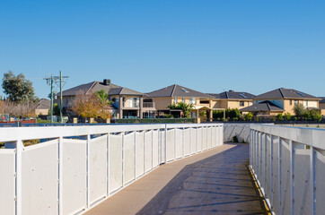 A pedestrian boardwalk leads to a residential neighbourhood with some two-storey modern houses. Sanctuary Lakes, Point Cook, Melbourne, VIC Australia.
