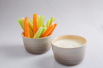 close up isolated shot of a mixed bowl of crunchy orange carrot slices and juicy green celery sticks with a white cup of blue cheese dipping sauce on a white background