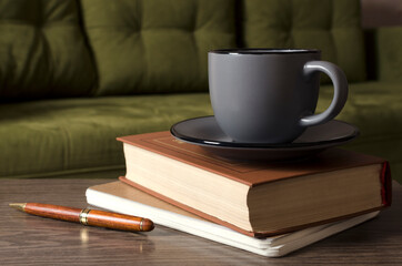 Closeup of cup of coffee on the stack of books, pen on the wooden table against green sofa.Empty space