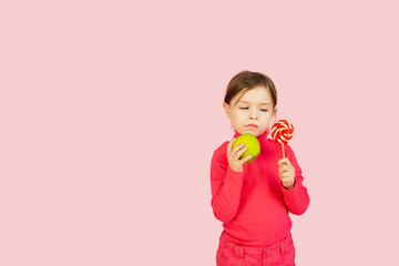 Little girl chooses between a lollipop and a green apple. The concept of proper nutrition. A child in a pink studio holds a sweet of sugar in his hand and an apple. Difficulty of choice