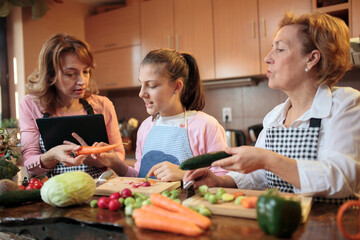 Beautiful teenage Caucasian girl preparing healthy vegetarian meal with her mother and grandmother. Mother is reading recipe on the tablet while grandmother is cutting cucumbers