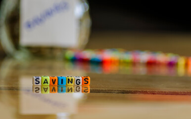 Conceptual image of savings. Only "savings" spelled colorful beads in focus. Reflective wooden surface. Others including other beads and coins-filled glass jar labeled "savings" in blur.