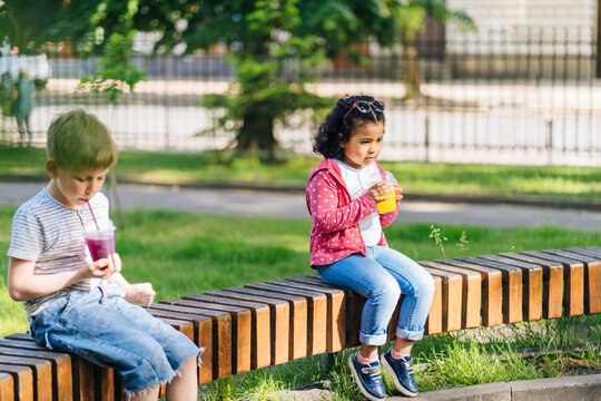Little Hispanic Preschooler Girl and Cute White Caucasian Boy Sitting On The Bench, drinking a fruit smoothie in summer city park.