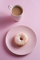 Vanilla donut with sprinkles on plate near to cup of coffee, sweet glazed dessert food and hot drink on pink minimal background, angle view