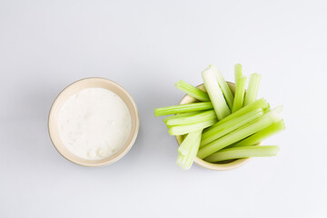 close up isolated flat lay top view shot of a bowl of crunchy juicy green celery sticks next to a white cup of blue cheese dipping sauce on a white background