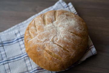 Round sourdough homemade bread with leaf scoring pattern on a wooden surface with a checked kitchen towel