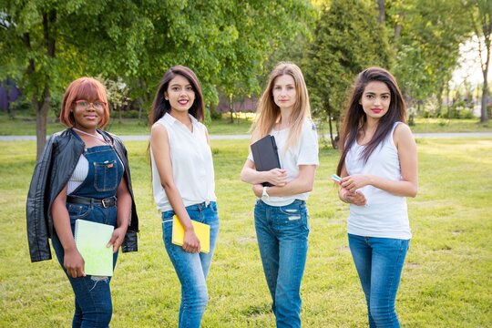 Four biracial female students or co-workers stand in the park. Women look at the camera, holding notebooks, books, tablets.