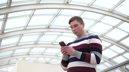 A young man is standing with a phone. A man uses a smartphone against a pan-glass roof. A busy person is texting in the messenger.