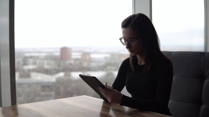 A young woman with a tablet sits at a table. A girl sits in a cafe by the panoramic window with a tablet in her hands. View from the window.