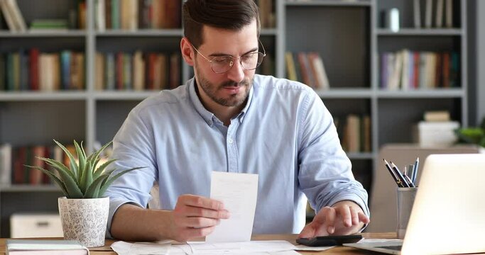 Focused young businessman in glasses holding paper receipt, doing accounting at office. Concentrated bearded man calculating household bills, managing monthly incomes and outcomes alone at home.