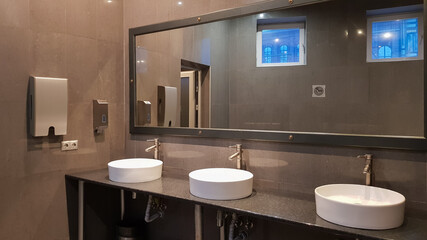 Chrome faucets with white round washbasins in a public toilet with a large mirror and gray walls,...