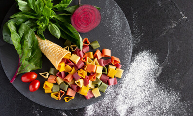 Colorful uncooked pasta in shape of heart on black slate board and fresh vegetables - basil, beetroot, cherry tomatoes, mangold leaves. Top view, copy space