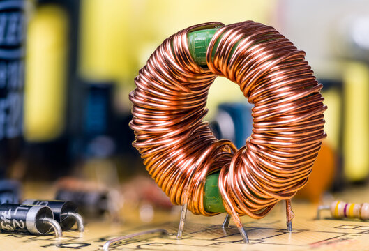 Induction coil with copper wire winding soldered on printed circuit board. Toroidal inductor with magnetic ferrite core. Electronic components on switch-mode power supply unit detail. Selective focus.
