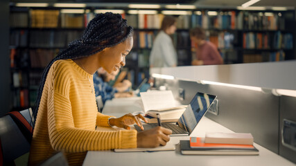 University Library: Gifted Beautiful Black Girl Sitting at the Desk, Uses Laptop, Writes Notes for...