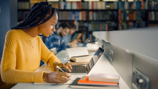 University Library: Gifted Black Girl Uses Laptop, Writes Notes For The Paper, Essay, Study For Class Assignment. Diverse Multi-Ethnic Group Of Students Learning, Studying For Exams, Talk In College