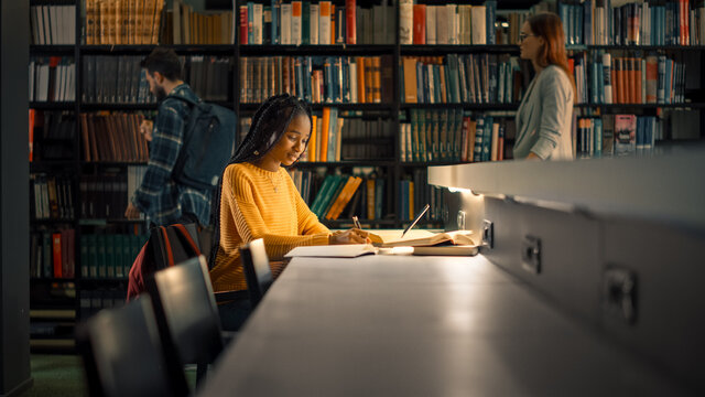 University Library: Gifted Black Girl uses Laptop, Writes Notes for the Paper, Essay, Study for Class Assignment. Students Learning, Studying for Exams College. Side View Portrait with Bookshelves