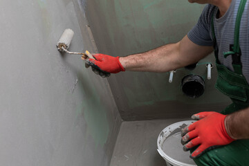 In the bathroom, a general construction worker applies moisture insulation using a roller.