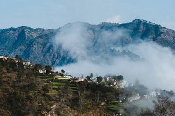 Fototapeta premium Aerial/Drone View of a foggy Himalayan valley and green fields surrounded by lush green hills during a beautiful morning at a rural location in Uttarakhand, India.