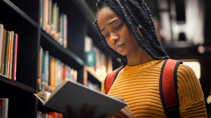 University Library: Smart Beautiful Black Girl Standing Next to Bookshelf Holding and Reading Text...