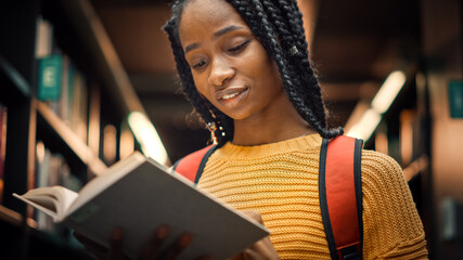 University Library: Smart Beautiful Black Girl Standing Next to Bookshelf Holding and Reading Text...