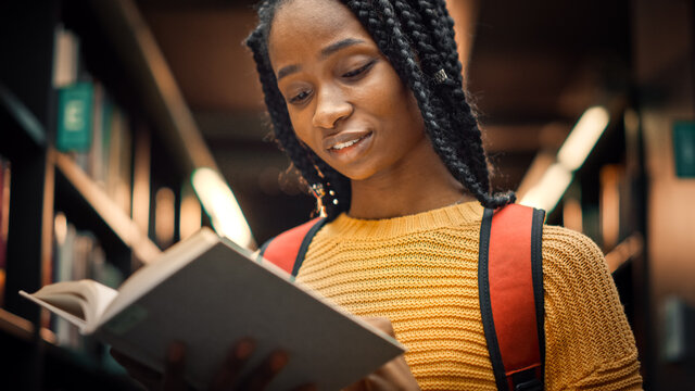 University Library: Smart Beautiful Black Girl Standing Next to Bookshelf Holding and Reading Text Book, Doing Research for Her Class Assignment and Exam Preparations. Low Angle Portrait with a Smile