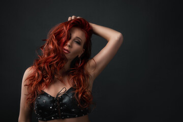Fototapeta premium concept of hair coloration and skin care with cosmetics, copy space. sexy girl touching her red colored hair. woman with professional make up wearing leather top bra with sparkling stars.