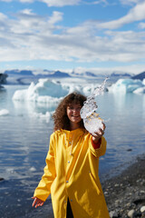 Must visit destination. Beautiful happy female tourist in yellow jacket showing crystal ice piece at camera and smiling while visiting Jokulsarlon glacier lagoon in Iceland