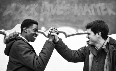 Black and white image of the joined hands of two young men of different ethnicity smiling at each...