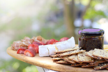 A cutting board with charcuterie. Spanish cured meat, prosciutto, brie cheese, crackers, jam and strawberries.