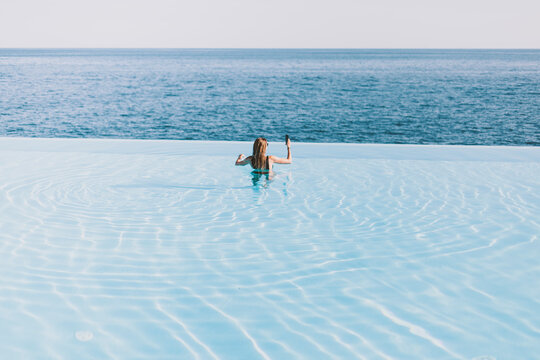 Girl takes pictures on the phone by the ocean while standing in the hotel’s infinity pool. Long hair, bikini.

