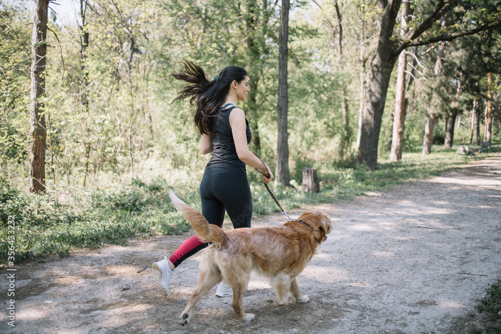 Wall mural Back view of girl and dog running in forest. Back of woman in sportswear running while holding leash of her dog in park.