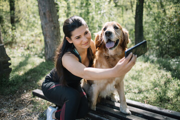 Beautiful girl taking photo with phone in forest. Young woman and her dog sitting on bench in nature while taking selfie.