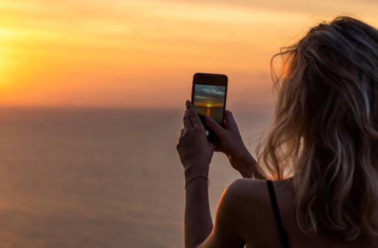 Woman hands holding mobile phone at sunset. Young curly hair woman taking photos with her cell phone in a beautiful amazing sunset over sea. Taking a picture on a smartphone during a vacation