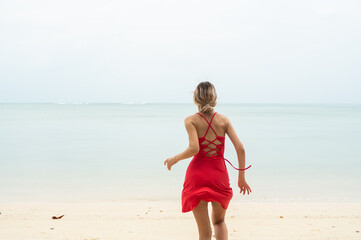 Young woman in a red dress runs and jumps on a white sand beach into tropical sea. Young happy woman running on the beach. Happiness to be on a sea or ocean. Vacation and travel concept.