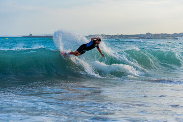 Spanish young man practicing skimboard on Mallorca's beaches with blue waves and clear sky with a black and blue wetsuit