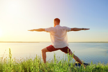 Fit muscular man doing yoga by the water on the grass with hands stretch. Orange sunlight sunset...