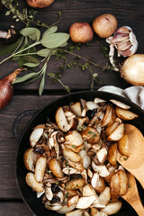 Roast forest mushrooms, potatoes, onions and seasonal herbs in a cast iron pan and ingredients on a wooden background outdoors.