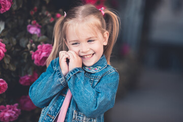 Smiling baby girl 3-4 year old laughing wearing denim jacket over rose flowers outdoors close up. Childhood. Summer time.